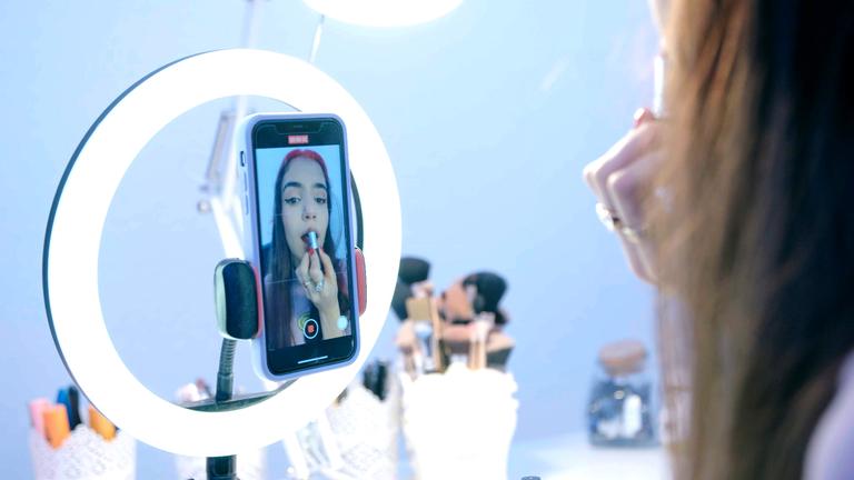  Die 17-jährige Hasti sieht sich in der Innenkamera ihres Handys an und schminkt sich dabei die Lippen. Das Handy ist an einem Ringlicht befestigt und auf einem Tisch befinden sich mehrere Make-up-Pinsel.