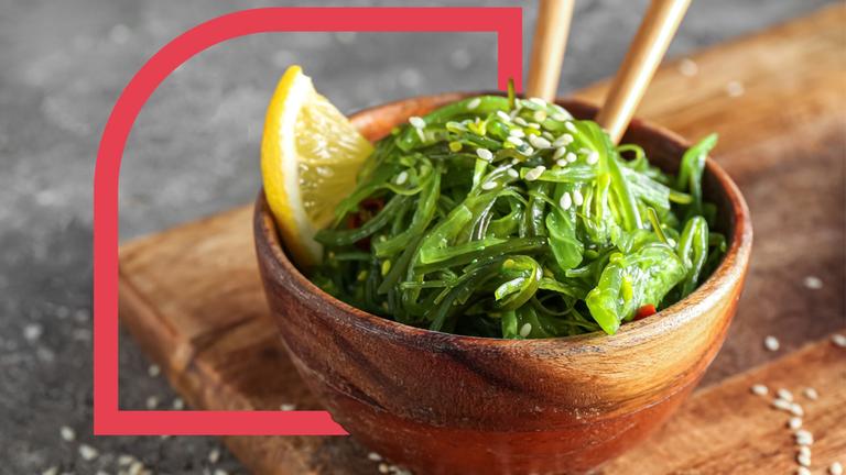 Eine Bowl mit Algen, mit Sesam bestreut und einer Zitronenscheibe garniert, in der Essstäbchen stecken.