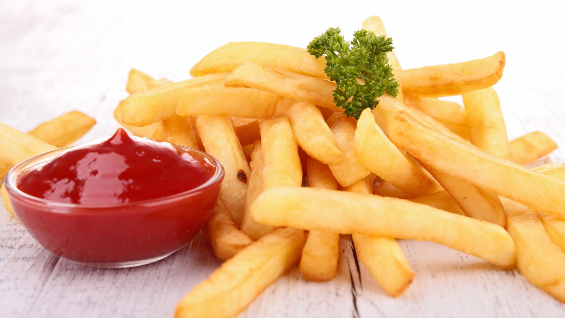 Eine Portion Pommes vor einem weißen Hintergrund. Als Garnitur liegt etwas krause Petersilie darauf. Daneben steht ein Glasschälchen mit Ketchup.