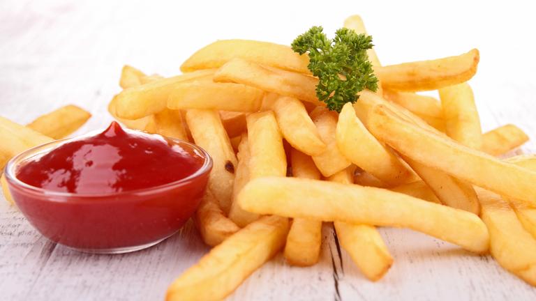 Eine Portion Pommes vor einem weißen Hintergrund. Als Garnitur liegt etwas krause Petersilie darauf. Daneben steht ein Glasschälchen mit Ketchup.