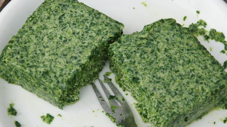 Ein Stück gefrorener Rahmspinat aus der Packung liegt von einer Gabel zerteilt auf einem Teller.
