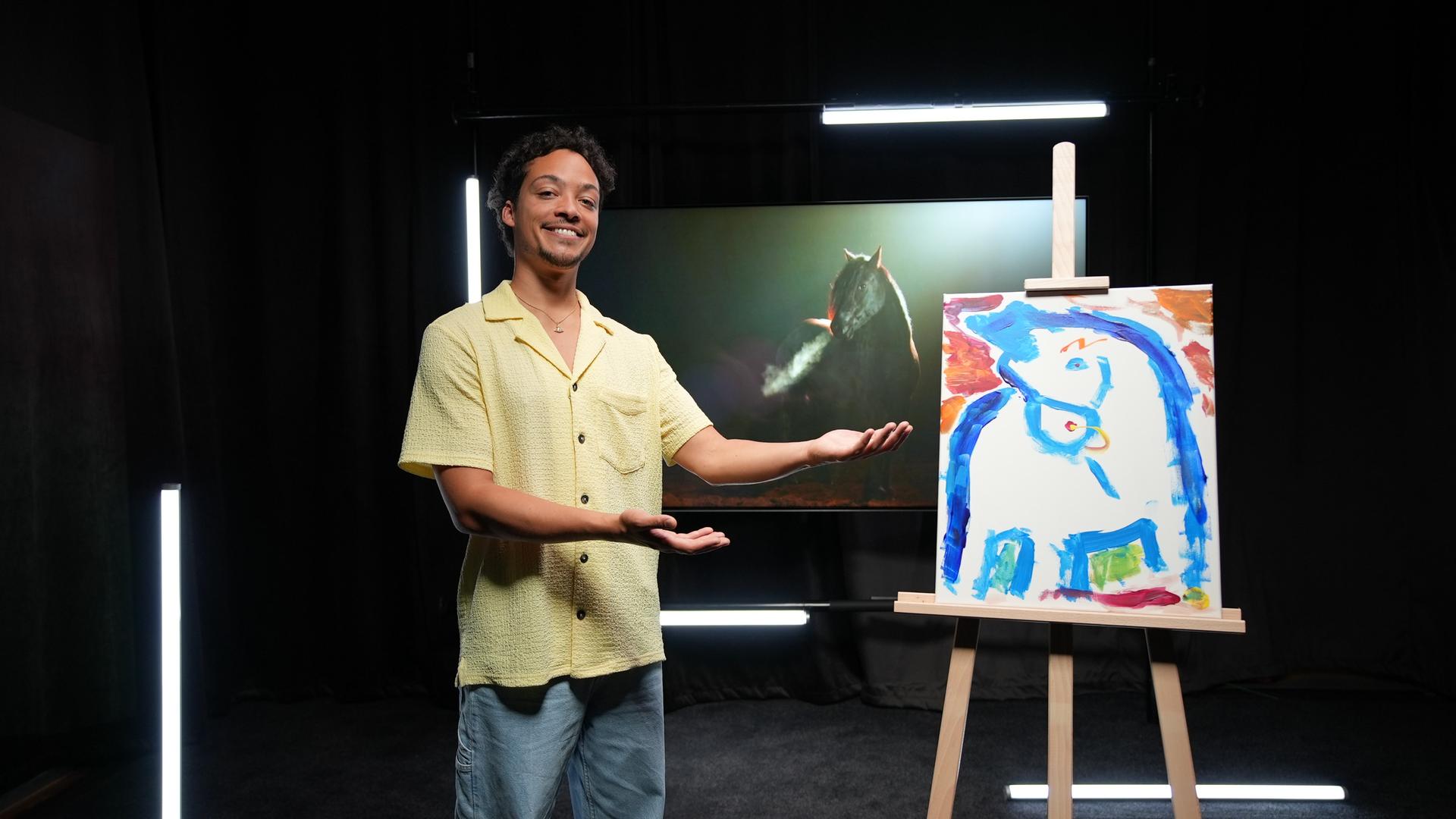 Ein Mann in gelbem Hemd präsentiert lächelnd ein Bild auf einer Staffelei. Drauf ist ein blaues Pferd zu sehen. Im Hintergrund sieht man ein großes Foto eines schwarzen Pferdes.