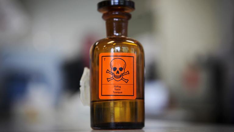 Braune Flasche mit orangenem Giftsymbol-Aufkleber steht auf einem Tisch.