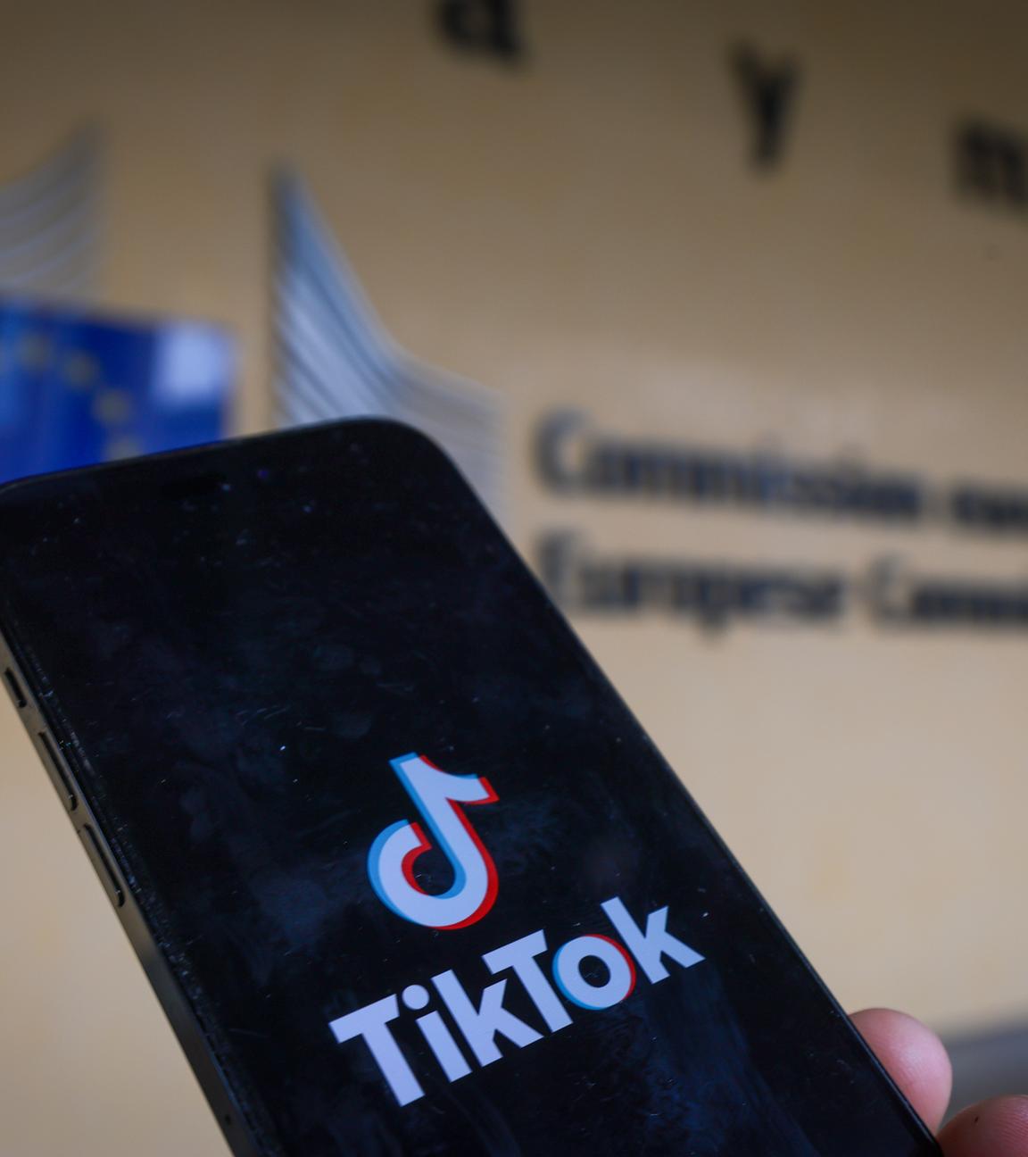 Ein Smartphone wird gehalten. Auf dem Bildschirm ist das Logo der Plattform Tiktok zu erkennen. Im Hintergund ist zu erkennen, dass es sich um den Hauptsitz der Europäischen Kommission in Brüssel, Belgien handelt. 