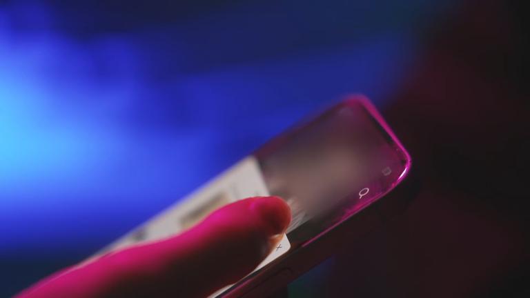 In der linken Bildhälfte ist eine Hand zu sehen, die ein Smartphone hält. Der Bildhintergrund ist blau und schwarz. Die Person scrollt womöglich durch soziale Medien