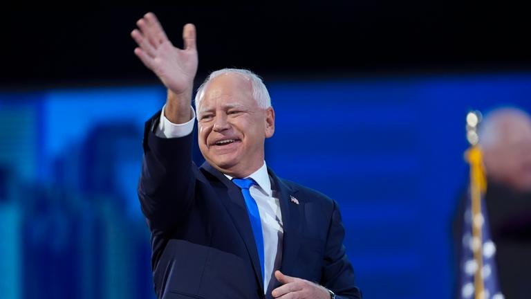 Tim Walz steht auf der Bühe, lächelt und winkt in die Menge.