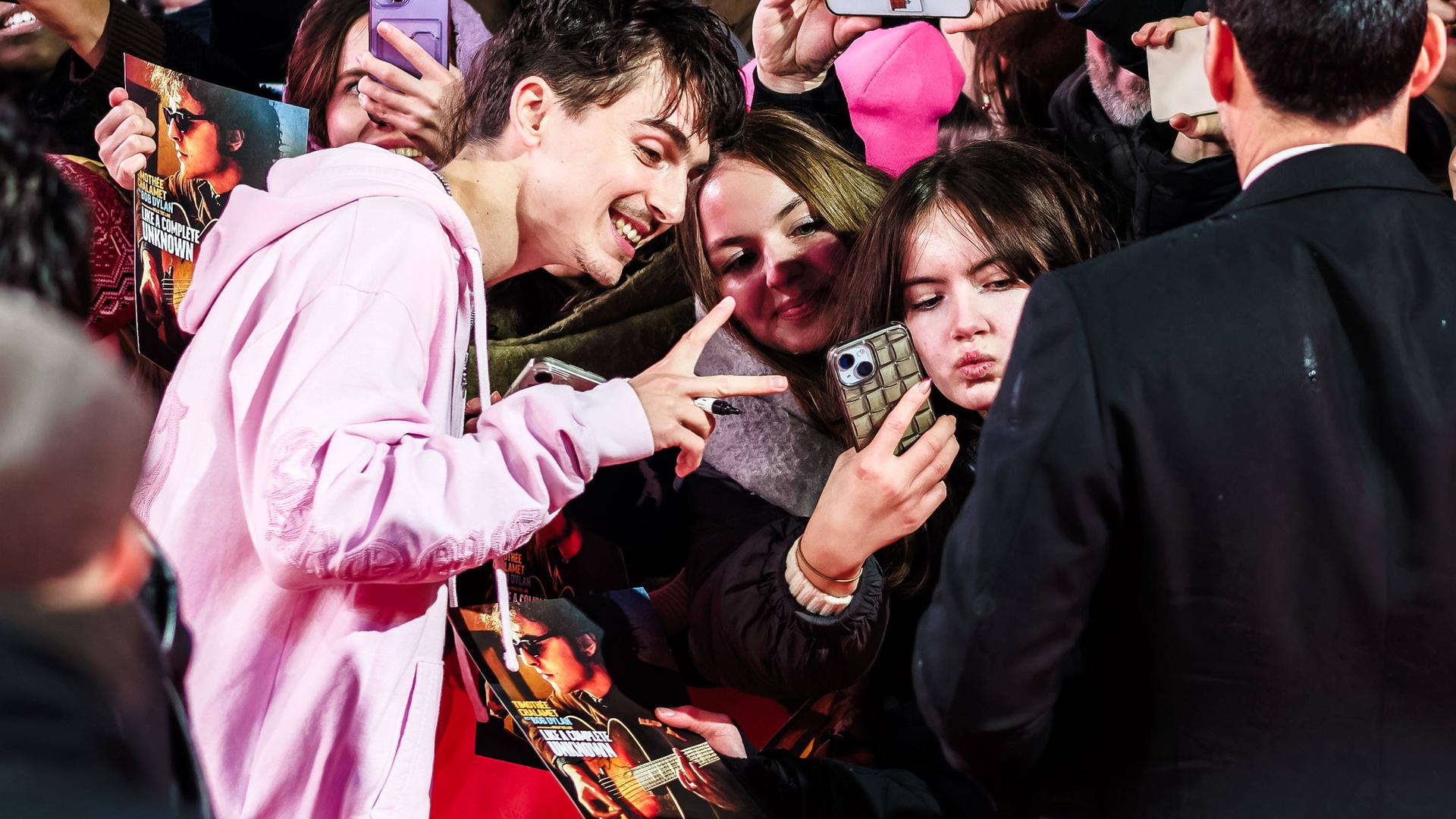 Ein junger Mann in einer rosa Sweatjacke zeigt seinen weiblichen Fans am roten Teppich der Berlinale das Vicotory-Zeichen und lächelt in die Handykameras.