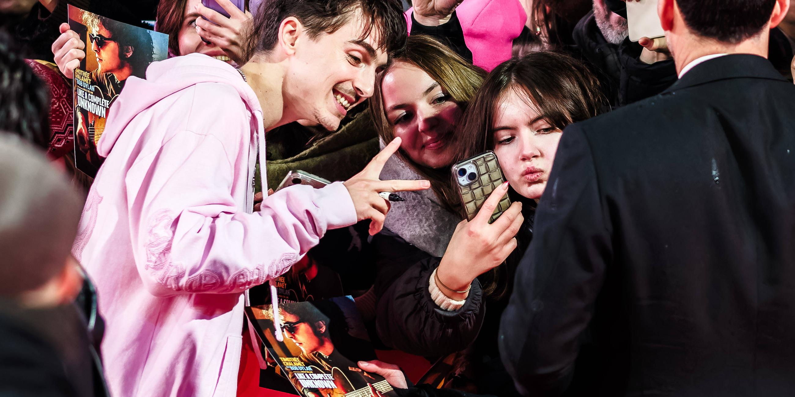 Ein junger Mann in einer rosa Sweatjacke zeigt seinen weiblichen Fans am roten Teppich der Berlinale das Vicotory-Zeichen und lächelt in die Handykameras.