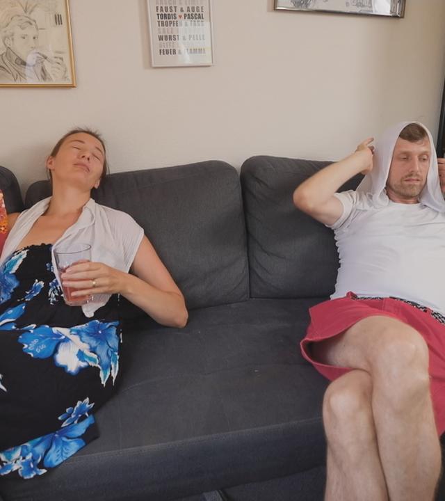 Ein Paar sitzt auf der Couch mit Fächer und Handtuch auf dem Kopf und will sich irgendwie abkühlen. 