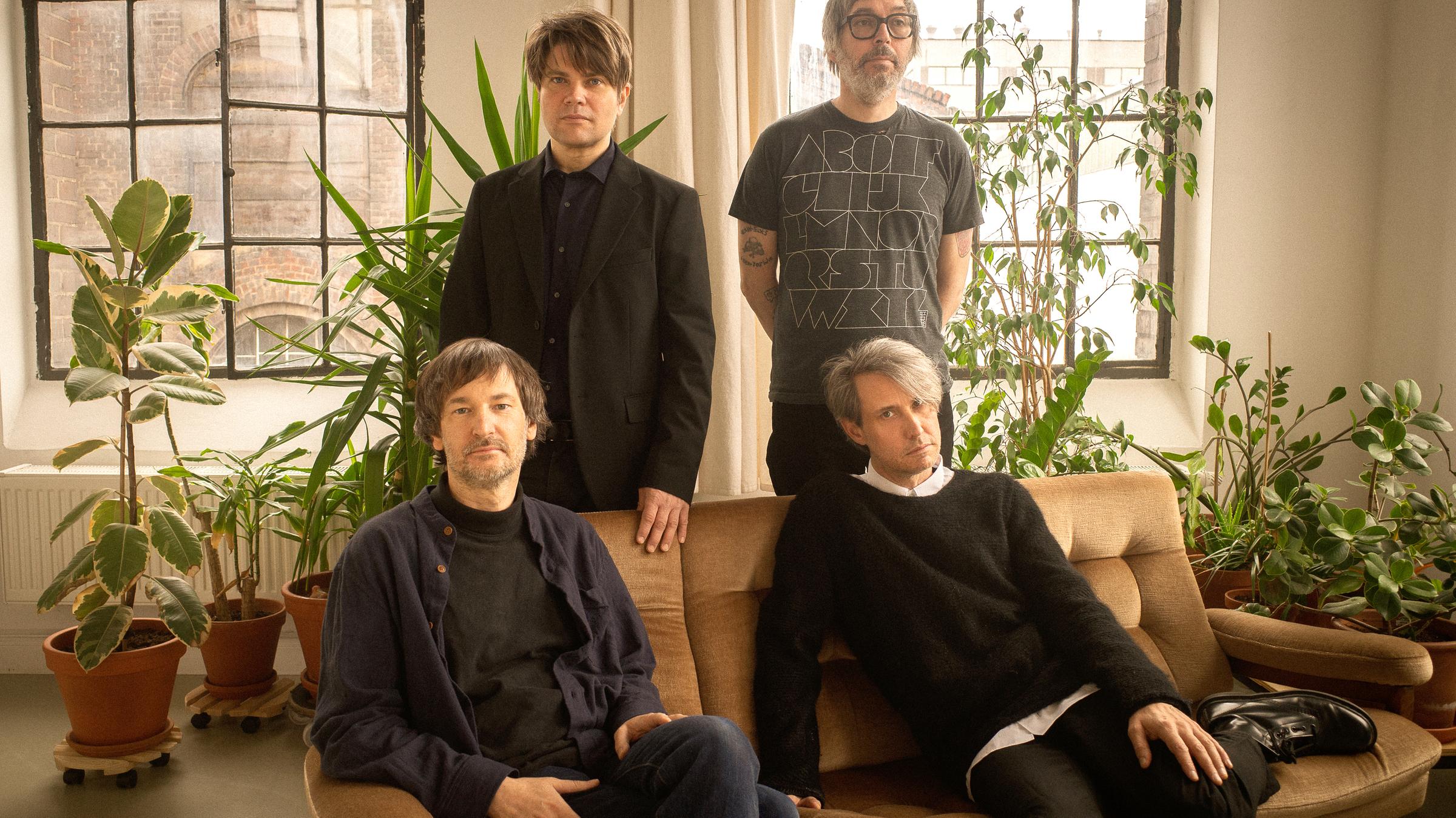 Zwei Männer in legerer Kleidung sitzen auf einem Sofa in einem mit Pflanzen bestücktem Raum. Hinter dem Sofa stehen zwei weitere Männer, die ebenfalls den Betrachter anvisieren.