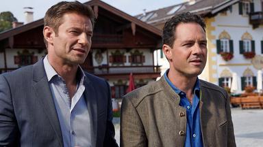 Die Kommissare Hansen (Igor Jeftić) und Aubinger (Thomas Birnstiel) stehen nebeneinander in einer Fußgängerzone und schauen in dieselbe Richtung. Im Hintergrund sind zwei bayerische Häuser mit Wandmalerei zu sehen.