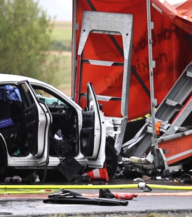 Das Bild zeigt einen beschädigten weißen Pkw nach einem Verkehrsunfall. Das Fahrzeug ist frontal in einen orangefarbenen Anhänger eines LKW gefahren.