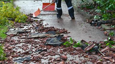 Personen räumen nach einem Tornado mit Schneeschiebern die zerstörten Dachziegel auf dem Gehweg vor einem Mehrfamilienhaus weg. 