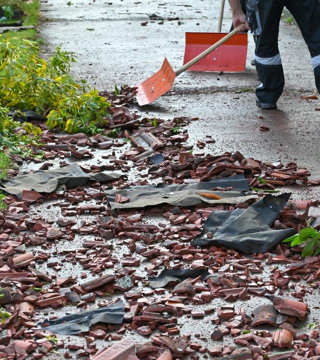 Personen räumen nach einem Tornado mit Schneeschiebern die zerstörten Dachziegel auf dem Gehweg vor einem Mehrfamilienhaus weg. 