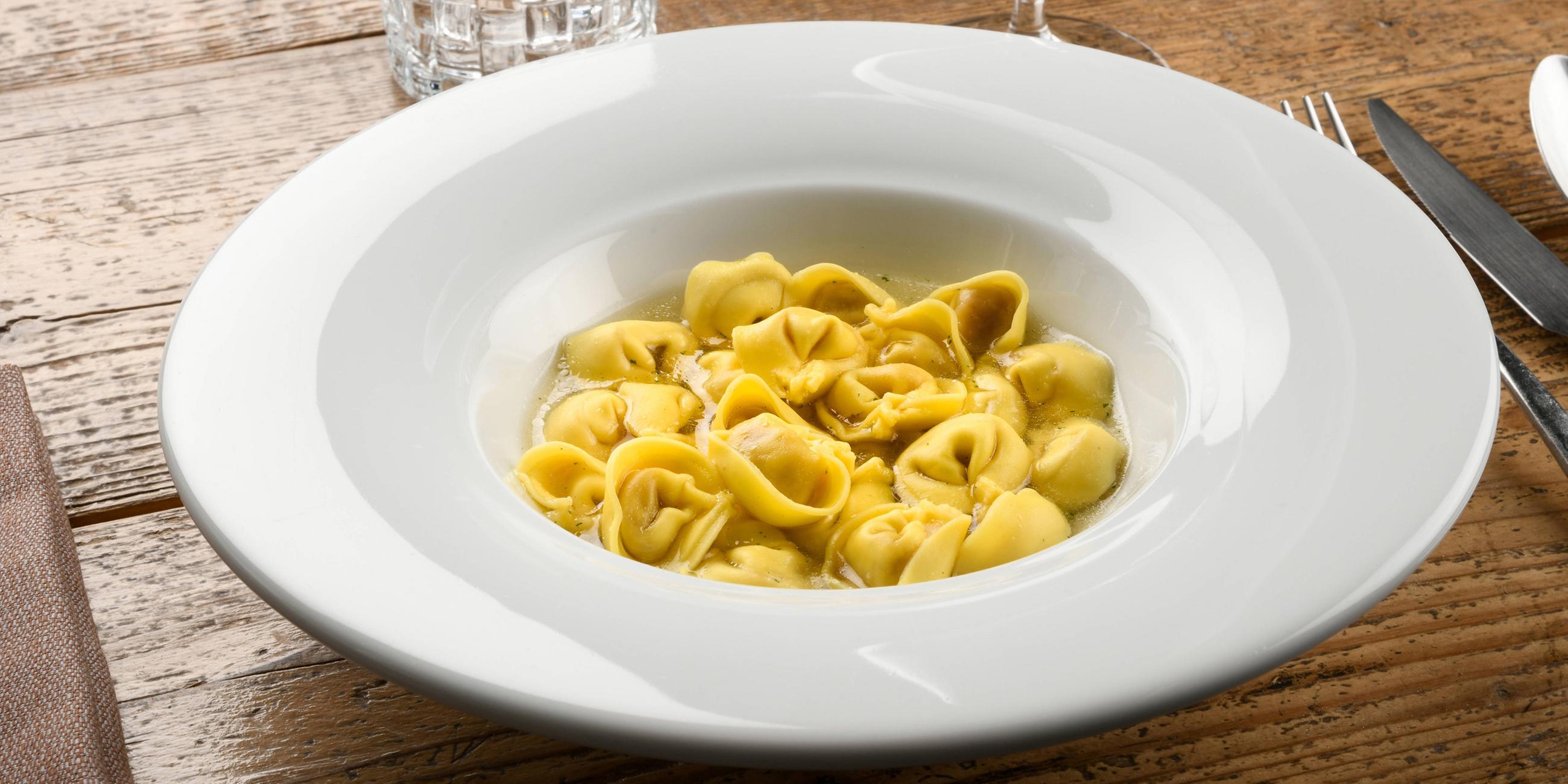 Schüssel mit Tortellini in Brühe aus der Region Emilia-Romagna in Italien, stilvoll in einem weißen Teller am Tisch serviert.