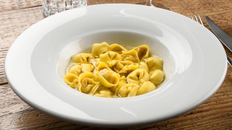 Schüssel mit Tortellini in Brühe aus der Region Emilia-Romagna in Italien, stilvoll in einem weißen Teller am Tisch serviert.