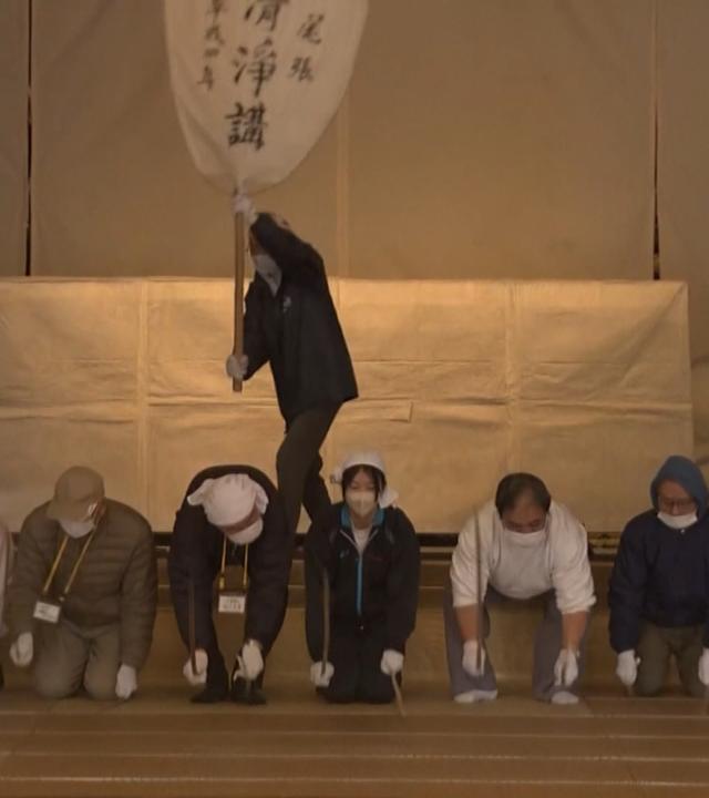 Menschen mit Atemschutzmaske knien auf dem Boden, im Hintergrund wedelt ein Mann mit einem großen weißen Fächer, auf dem schwarze Japanische Schriftzeichen sind.