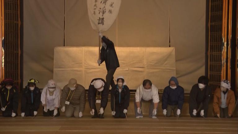 Menschen mit Atemschutzmaske knien auf dem Boden, im Hintergrund wedelt ein Mann mit einem großen weißen Fächer, auf dem schwarze Japanische Schriftzeichen sind.