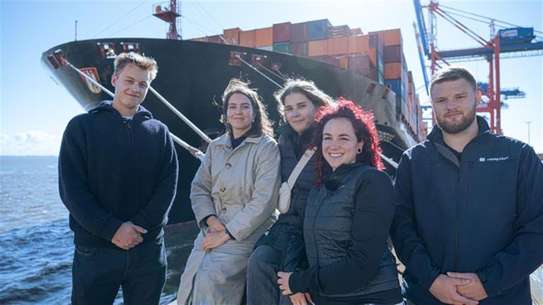 Ansgar Stark, Gweny Backs, Sofia Spirgatis, Selina Schwarz und Jamie von Bresinski im Hafen vor dem Containerschiff Chicago Express strahlen in die Kamera.