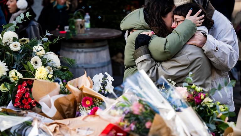 Menschen liegen sich am 02.01.2026 trauernd in den Armen vor niedergelegten Blumen, um den Opfern zu gedenken, die während der Neujahrsfeierlichkeiten in Crans-Montana durch einen Brand in der Schweizer Bar und Lounge „Le Constellation” ums Leben kamen und verletzt wurden. 