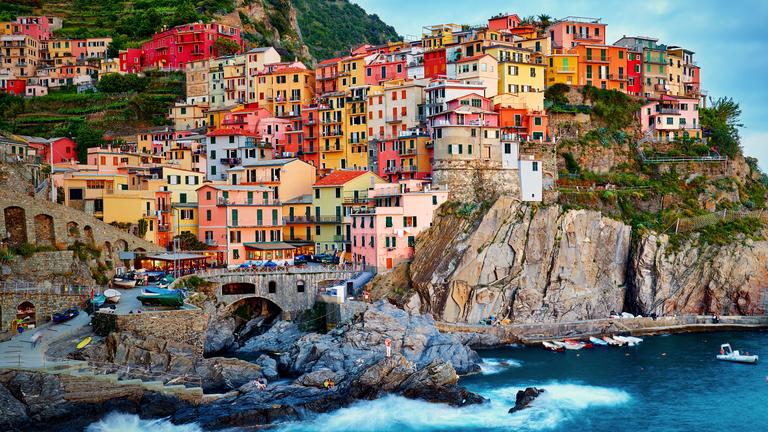 Blick vom Meer aus auf ein Dorf, auf Manarola. Bunte Häuser schmiegen sich an eine steile, felsige Küste.