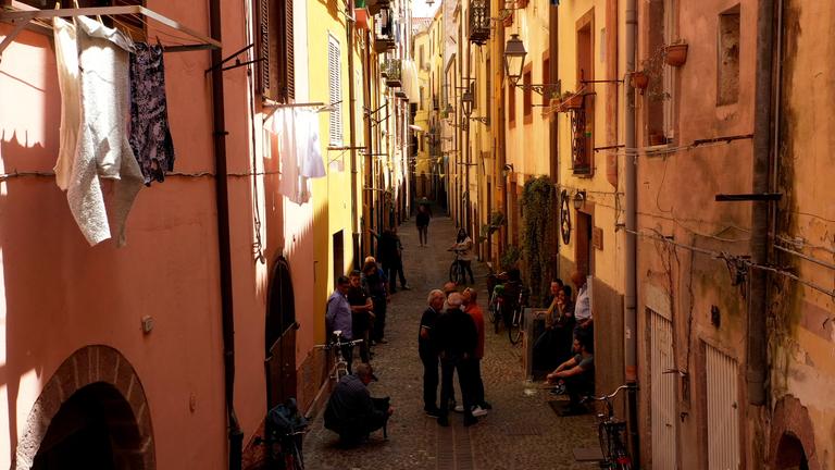 Enge Altstadtgasse im Dorf Bosa mit ockergelben Häuserwänden und einigen Menschen.