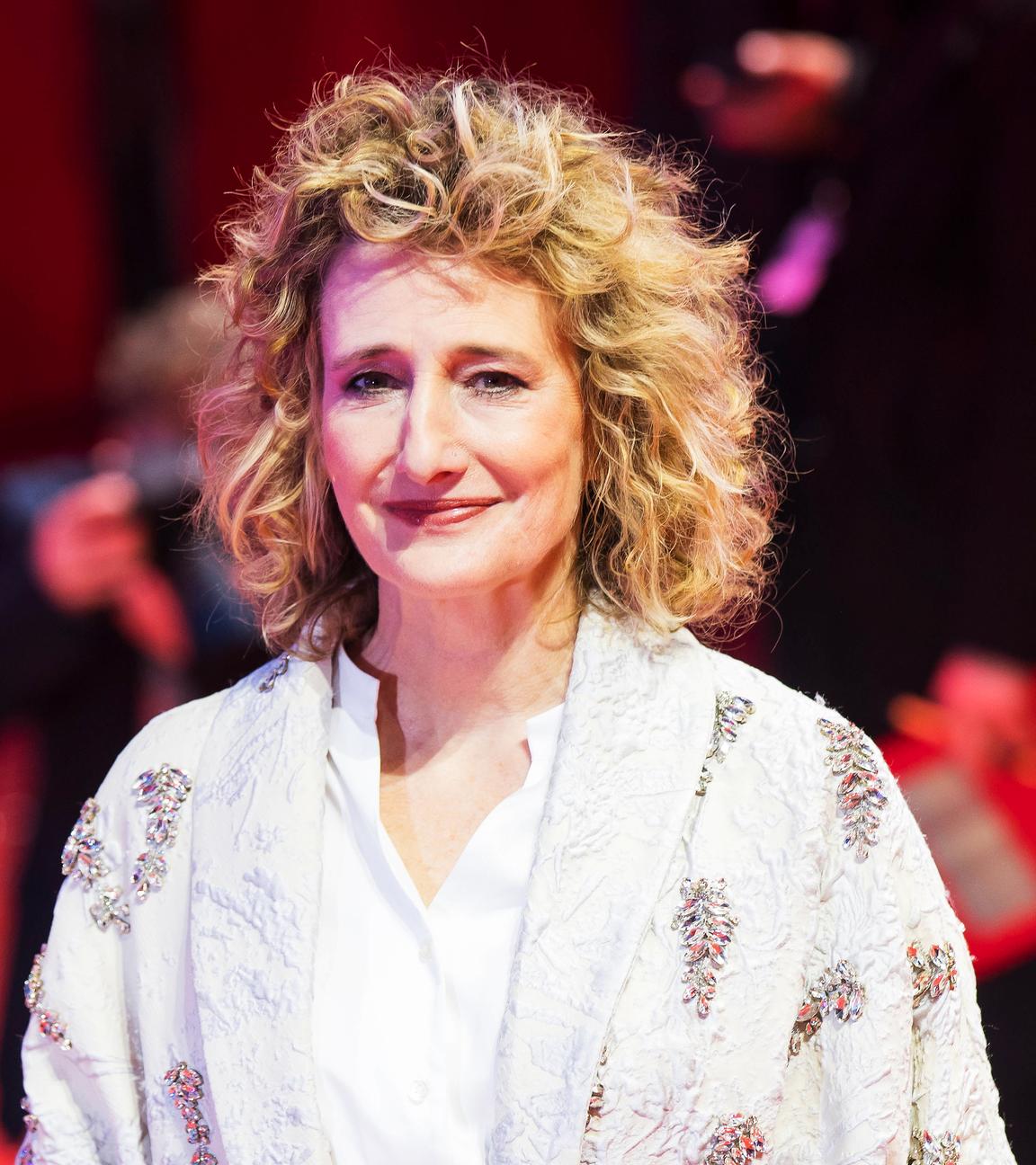 Eine Frau (Tricia Tuttle) mit blonden lockigen Haaren, die eine Bluse und einen Mantel trägt. Sie steht auf einem roten Teppich und hinter ihr sind Fotografen.