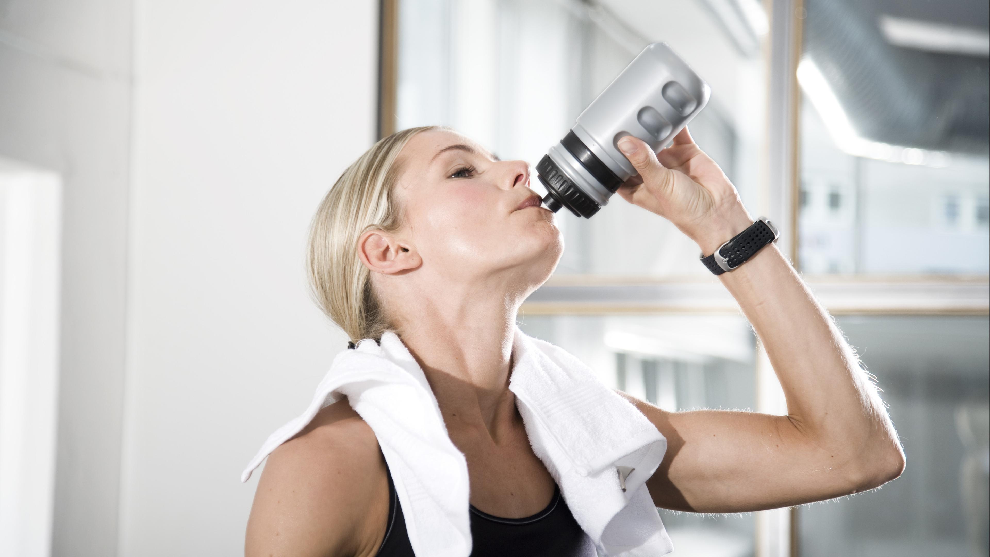Eine Frau hat ein weißes Handtuch über den Schultern liegen und neigt ihren Kopf nach hinten um aus einer grauen Plastikflasche zu trinken.