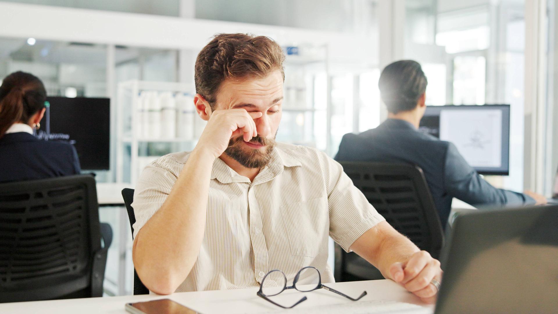 Ein Mann, der im Büro an einem Laptop arbeitet, reibt sich die Augen.