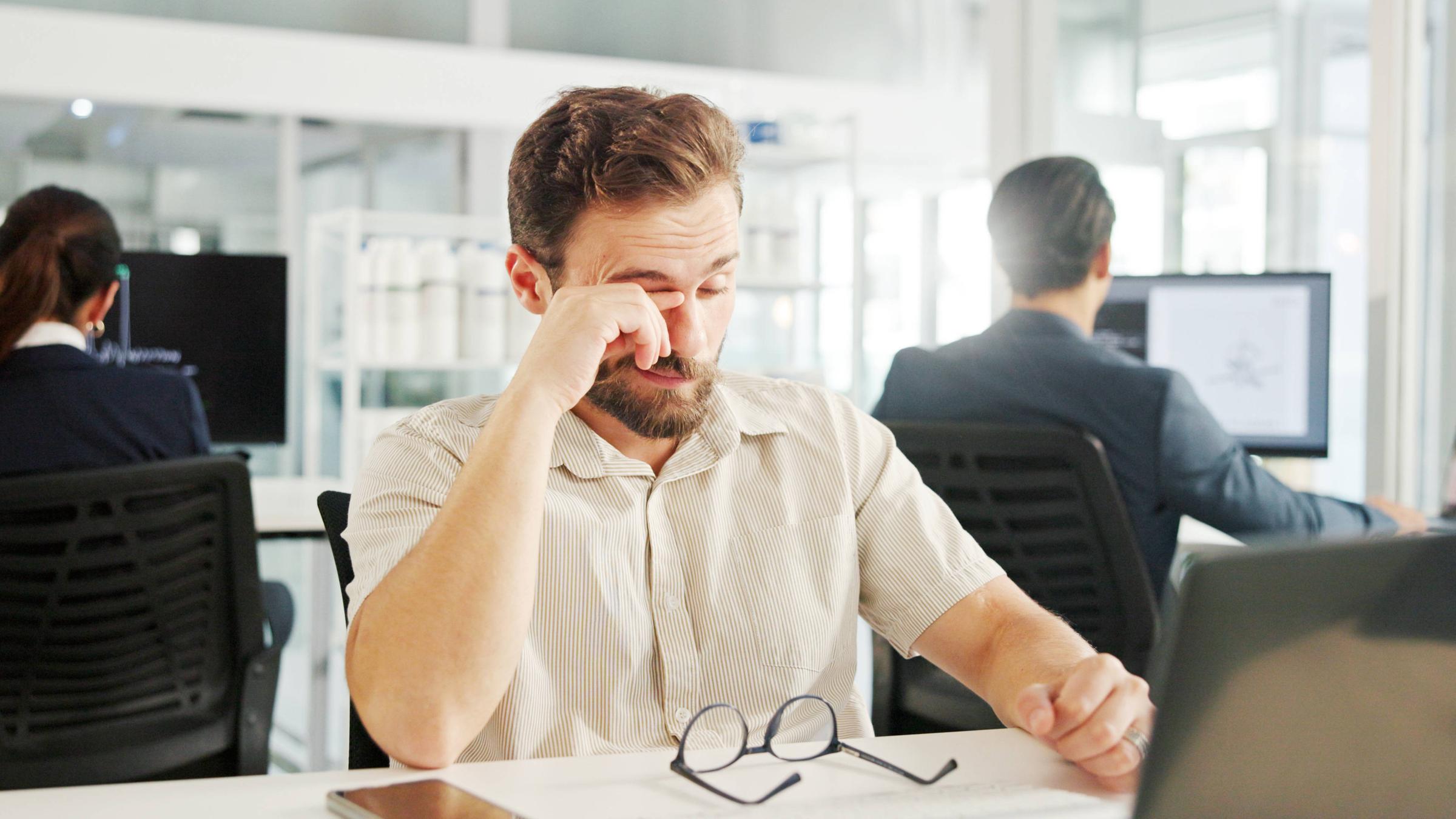 Ein Mann, der im Büro an einem Laptop arbeitet, reibt sich die Augen.