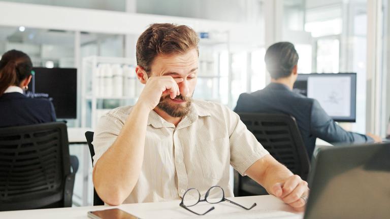 Ein Mann, der im Büro an einem Laptop arbeitet, reibt sich die Augen.