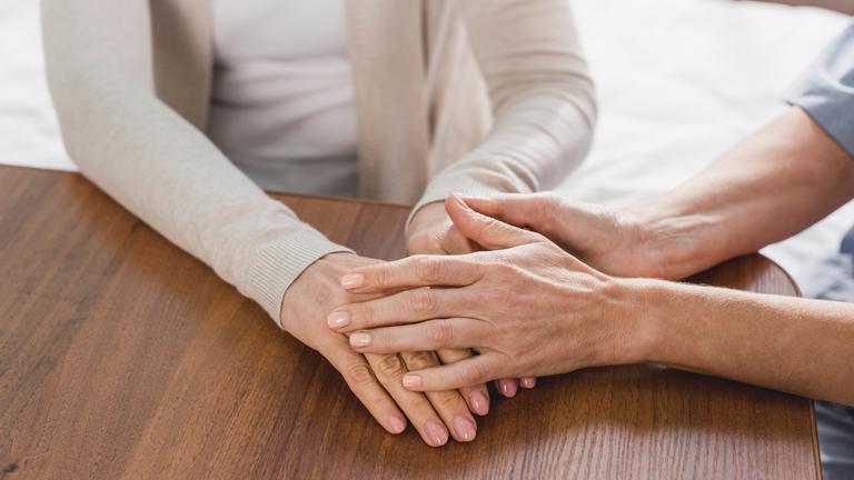Zwei Menschen sitzen an einem Tisch. Der Mann legt der Frau behutsam seine Hände auf ihre. 