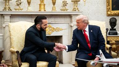 US-Präsident Donald Trump und der Präsident von El Salvador, Nayib Bukele, sitzen in Anzügen in hell gepolsterten schweren Sesseln vor einem mit goldenen Pokalen geschmückten Kamin im Weißen Haus und geben sich die Hand.