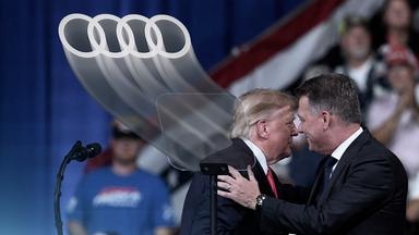 Links sind die typischen vier Audi-Ringe zu sehen. Rechts umarmen sich Donald Trump und Audi-Chef Gernot Döllner.
