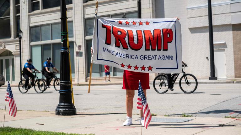 Eine Frau im roten Kleid hebt eine Trump-Flagge in die Höhe, sodass ihr Kopf verdeckt ist. Hinter ihr fahren Polizisten auf dem Fahrrad vorbei.