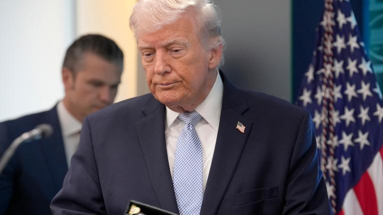 US-Präsident Donald Trump im Presseraum des Weißen Hauses mit gesenktem Blick. Im Hintergrund steht Kriegsminister Hegseth.