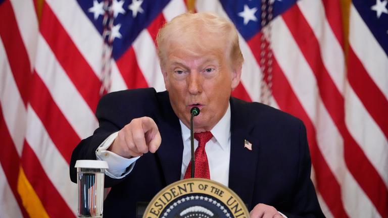 US-Präsident Trump spricht am Mikrofon vor mehreren USA-Flaggen und zeigt mit dem Zeigefinger an der Kamera vorbei.