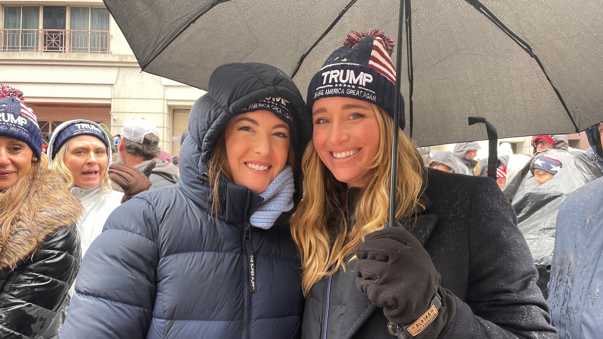 Zwei junge Frauen stehen unter einem Regenschirm und lächeln in die Kamera.