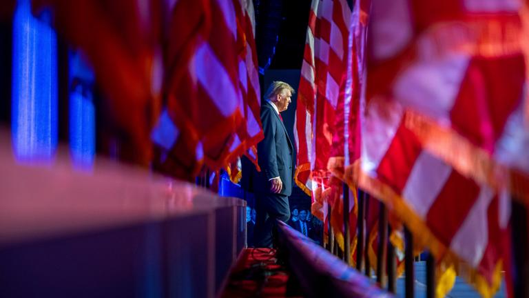 Zwischen mehreren US-Flaggen sieht man Donald Trump auf der Bühne stehen.