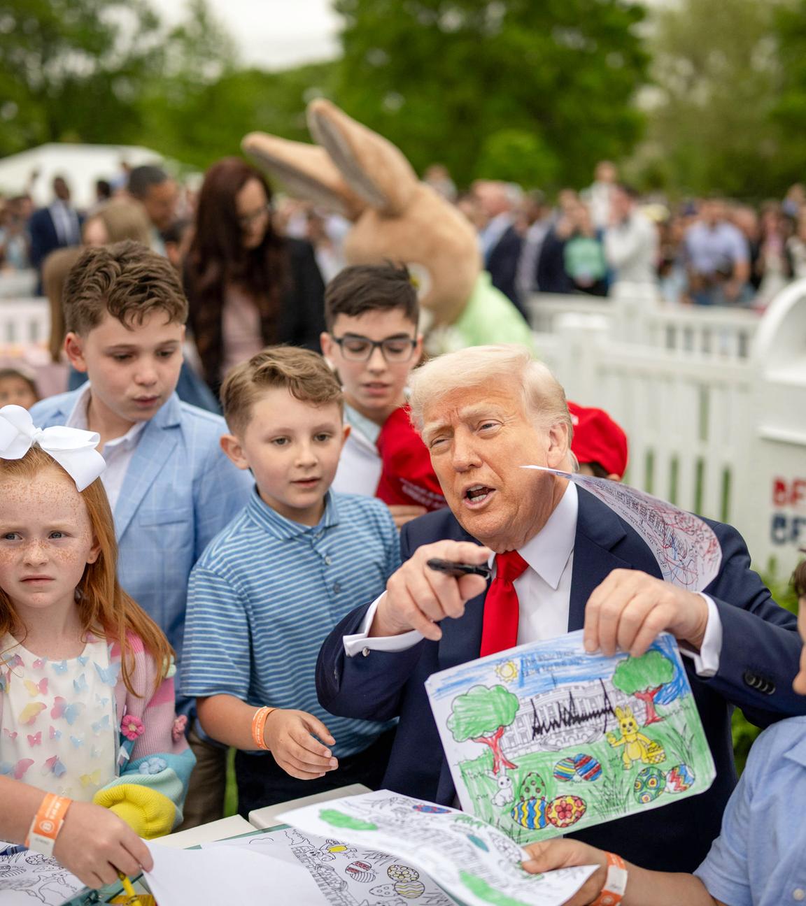 Ein älterer Mann im Anzug sitzt an einem Tisch, umringt von Kindern und hält ein gemaltes Bild und einen Stift in den Händen.