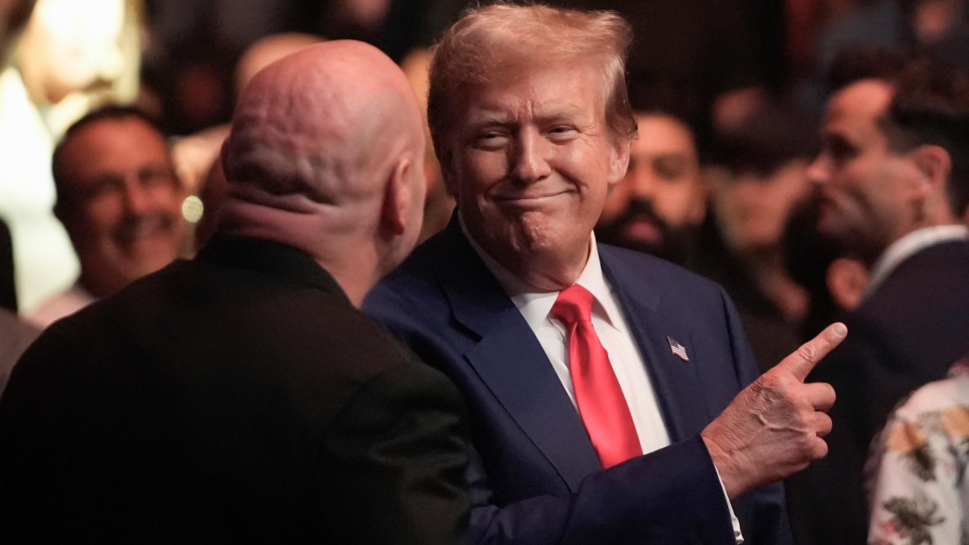 Donald Trump steht im blauen Anzug mit roter Krawatte in einer Menge und zeigt mit dem Zeigefinger nach oben.