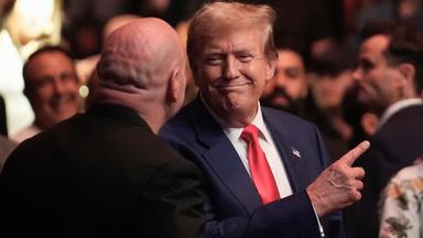 Donald Trump steht im blauen Anzug mit roter Krawatte in einer Menge und zeigt mit dem Zeigefinger nach oben.