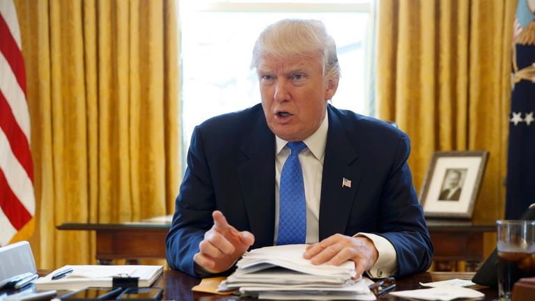 Donald Trump sitzt im Oval Office hinter dem großen Schreibtisch und zeigt mit erhobenem Zeigefinger in Richtung Kamera.