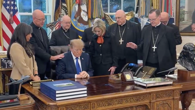 Donald Trump sitzt an seinem Schreibtisch im Oval Office. Um ihn herum stehen katholische Bischöfe, die mit ihm beten.