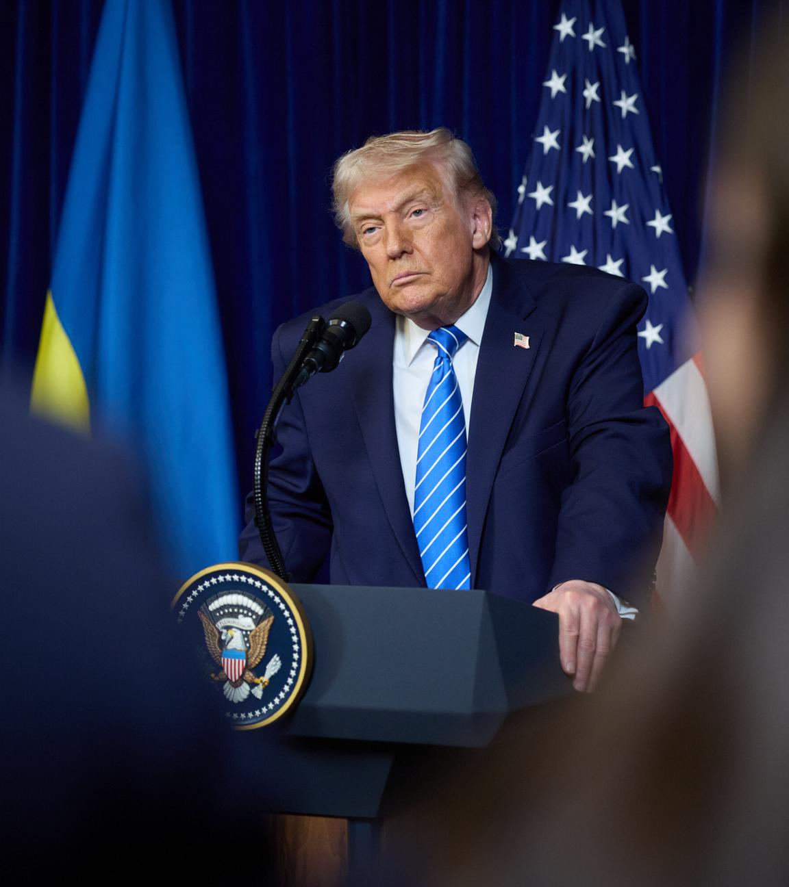 Donald Trump steht am Rednerpult mit ernstem Blick. Hinter ihm die US-Flagge und doe ukrainische Flagge.