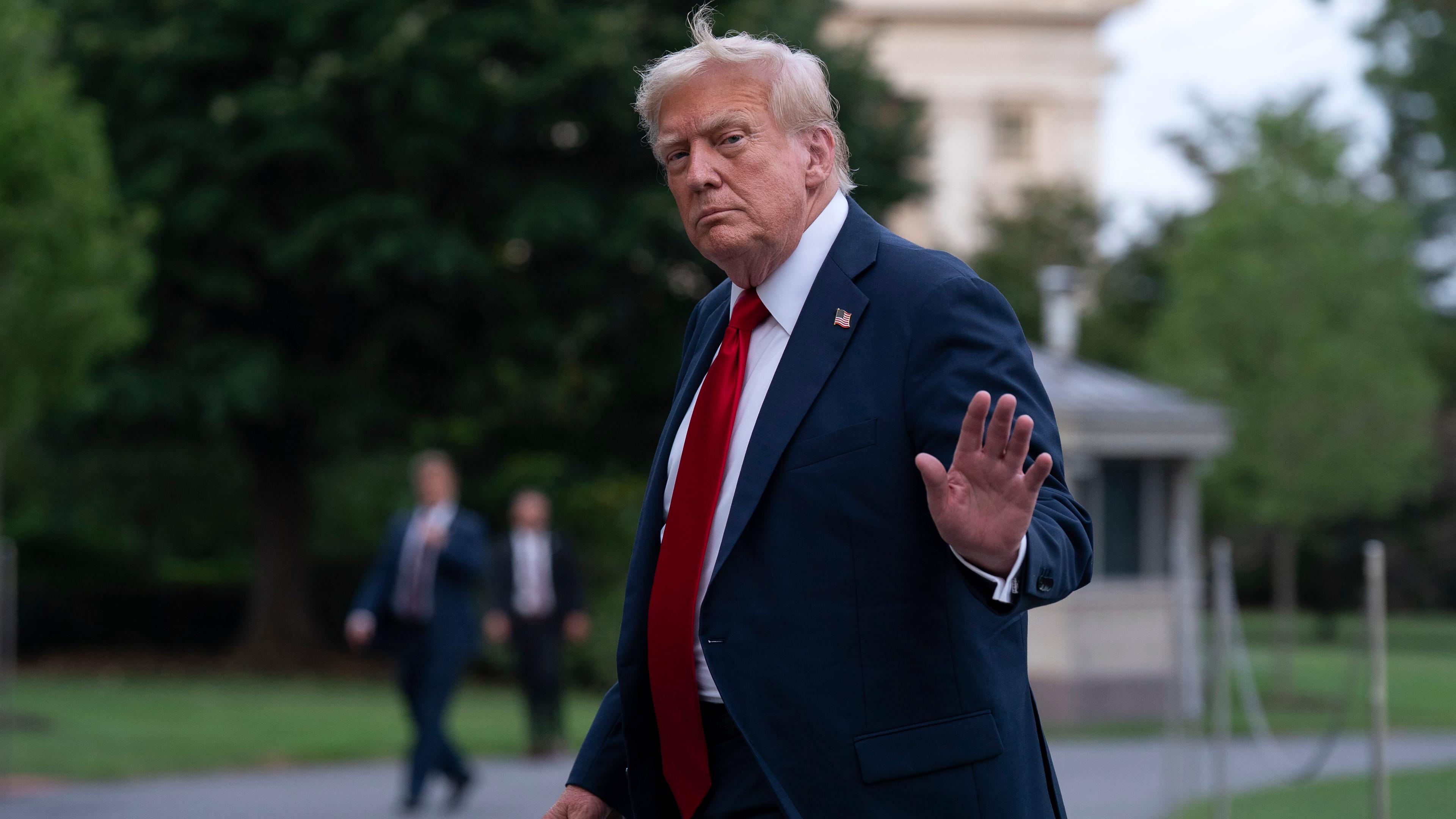 US-Präsident Trump trägt einen dunklen Anzug mit Krawatte und einer Stecknadel mit der Flagge der USA. Er winkt den Reportern.