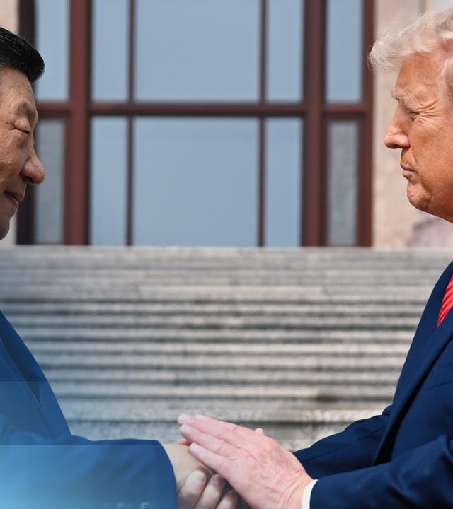 Das Seitenprofil von Trump und Xi. Sie stehen sich gegenüber. Xi lächelt leicht, Trump guckt eher ernst. Sie tragen beide einen blauen Anzug und schütteln sich die Hand.