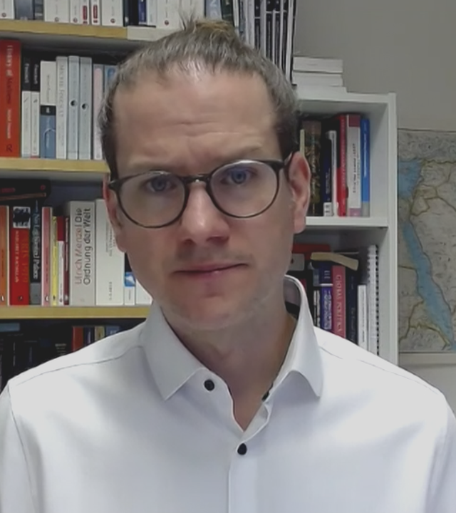 Nahost-Experte Jan Busse live zugeschaltet per Video-Call aus einem Büro mit einem vollen Bücherregal hinter sich.