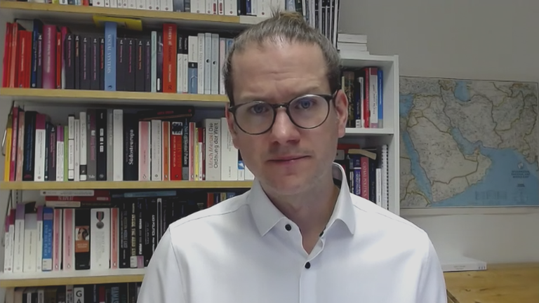 Nahost-Experte Jan Busse live zugeschaltet per Video-Call aus einem Büro mit einem vollen Bücherregal hinter sich.
