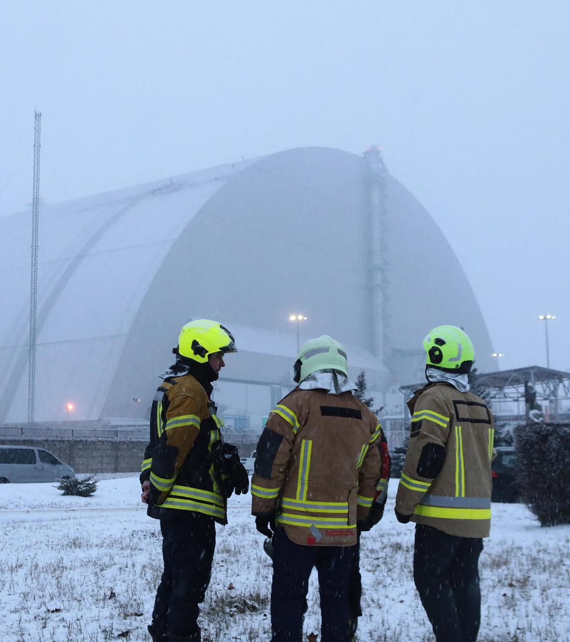 Drei Feuerwehrmänner stehen vor der zum Teil zerstörten Schutzhülle des Kraftwerks in Tschernobyl, welche im Januar 2025 durch eine russische Drohne angegriffen wurde.